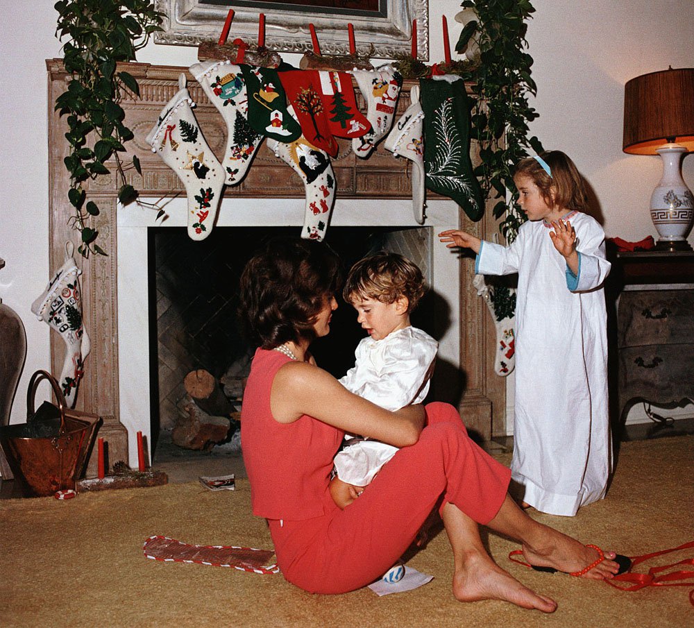 Jacqueline Kennedy with the children on Christmas morning. She sits on the floor, holding John on her lap, and Caroline stands nearby in her nightgown. Several stockings hang from the mantle.