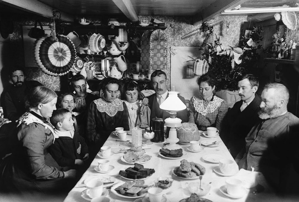 Men, women, and children assembled around a table spread with cakes, biscuits, and crockery.