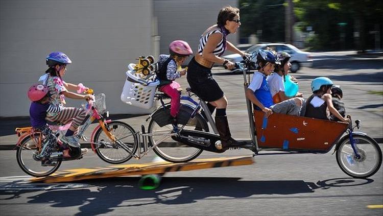 random picture of of woman riding bike contraption with 7 kids hanging on along with toys and going over some kind of plank