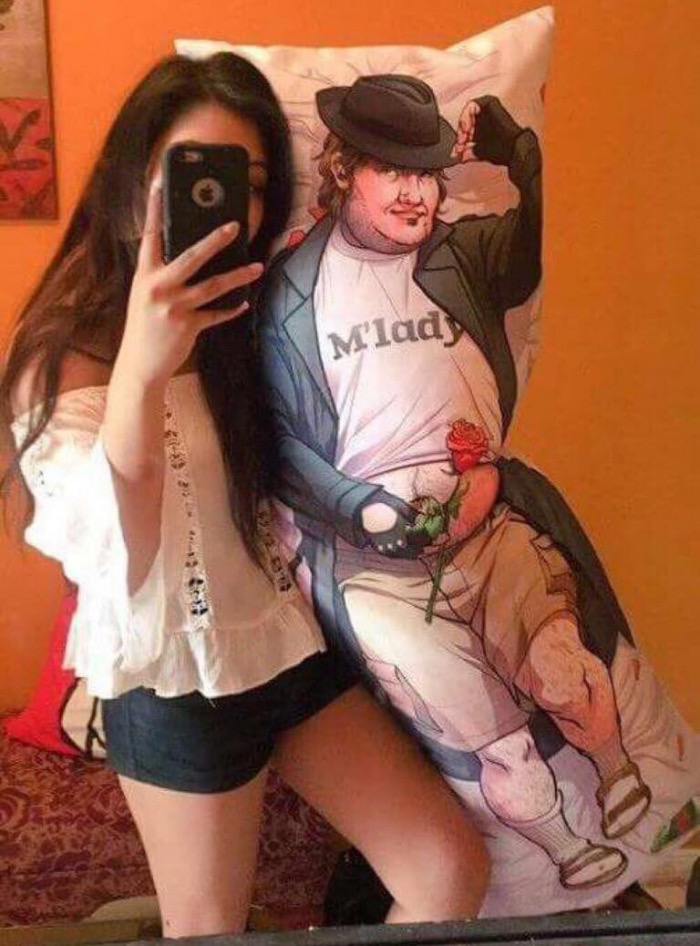 Awesome pic of a woman with a waifu pillow of a man with fedora, neckbeard, t-shirt that says m'lady, holding a rose and wearing cargo shorts with trenchcoat but socks with sandles.