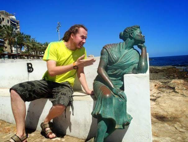 man posing with greenish copper statue of woman as if she is angry at him and avoiding conversation