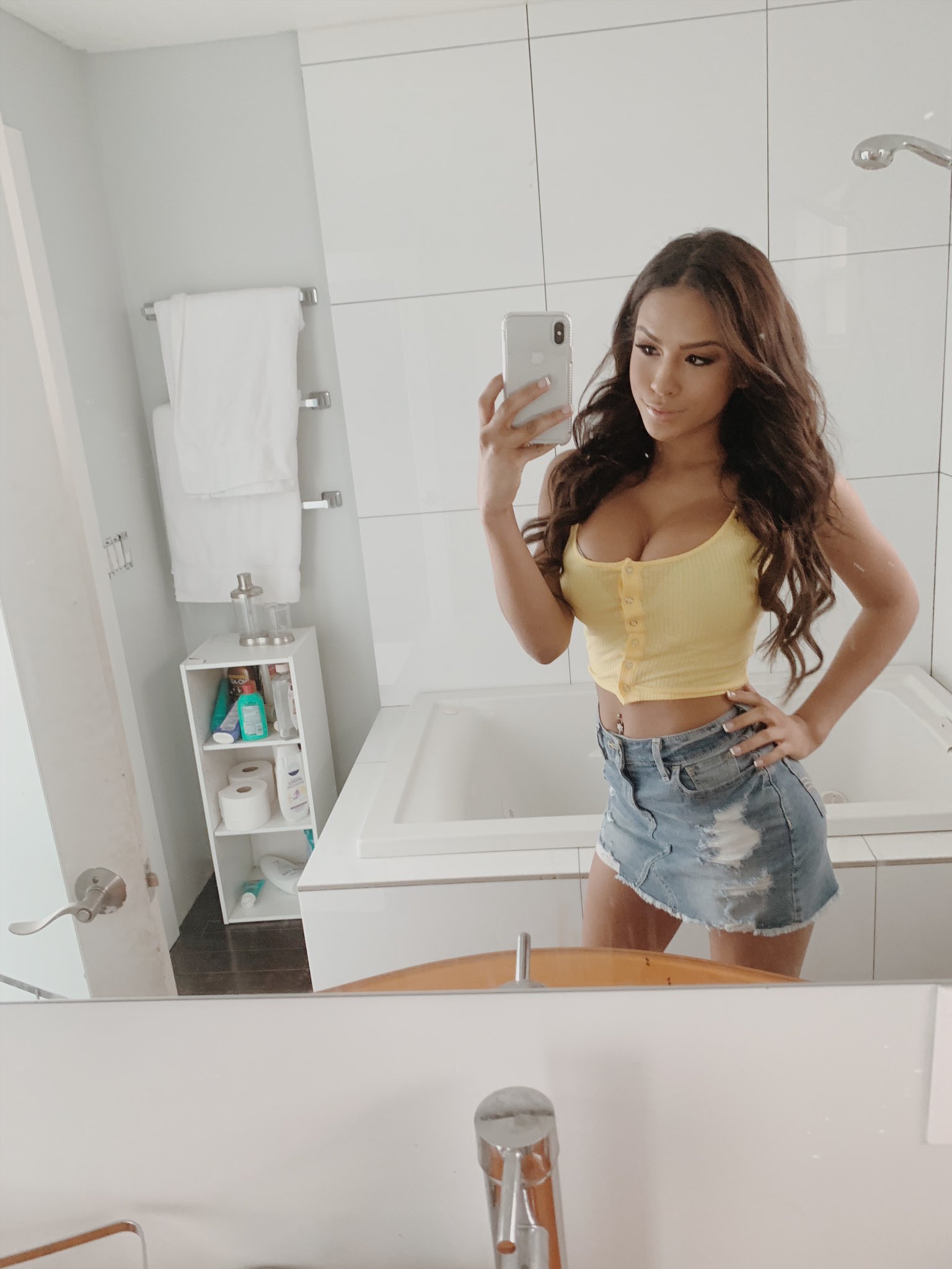 Sexy porn star Autumn Falls taking a mirror selfie in a bathroom wearing a tight pink shirt and a jean skirt.