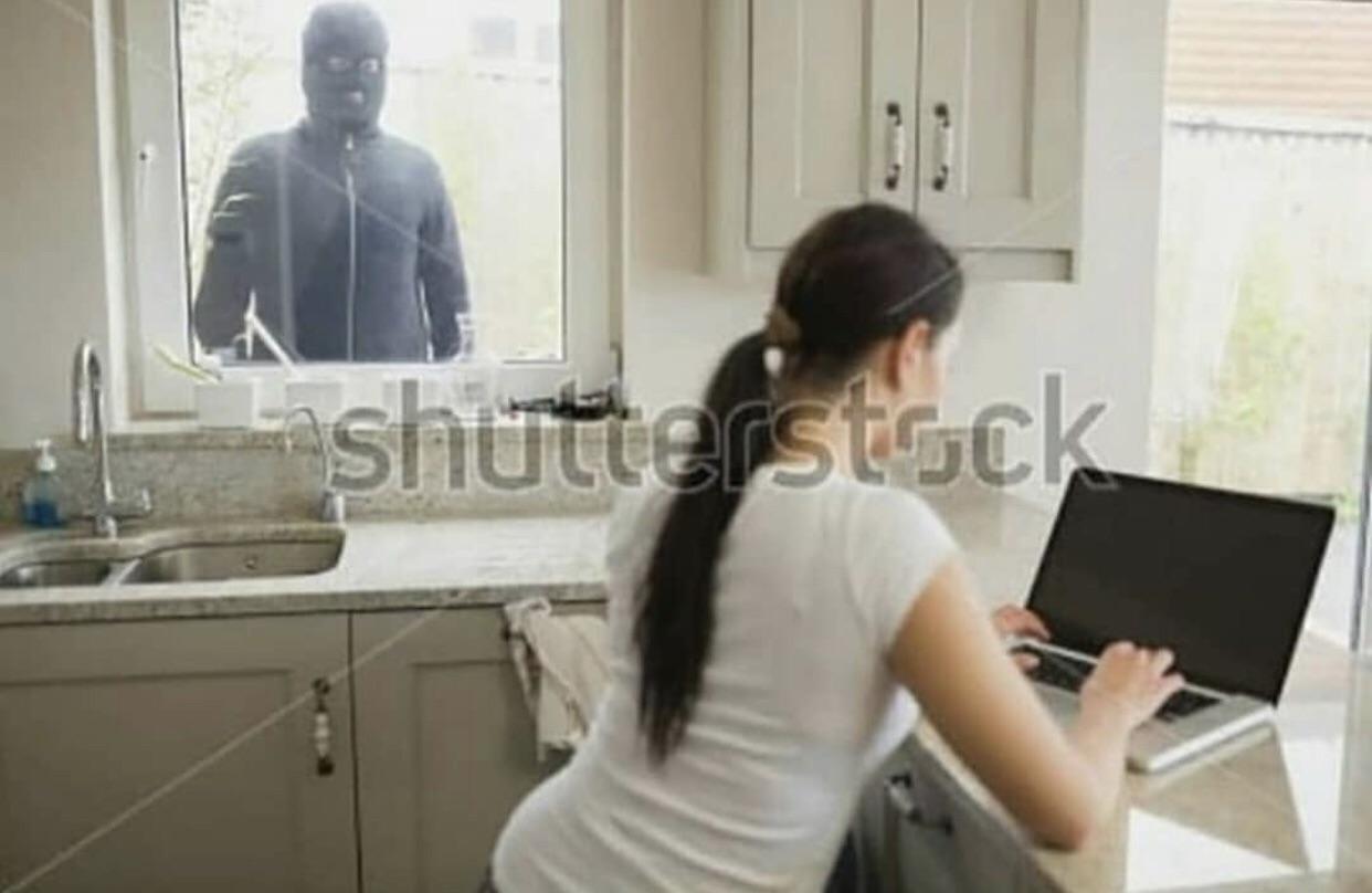 A funny and disturbing stock photo of a burglar standing outside a window obviously staring at a woman in her kitchen on a computer. 