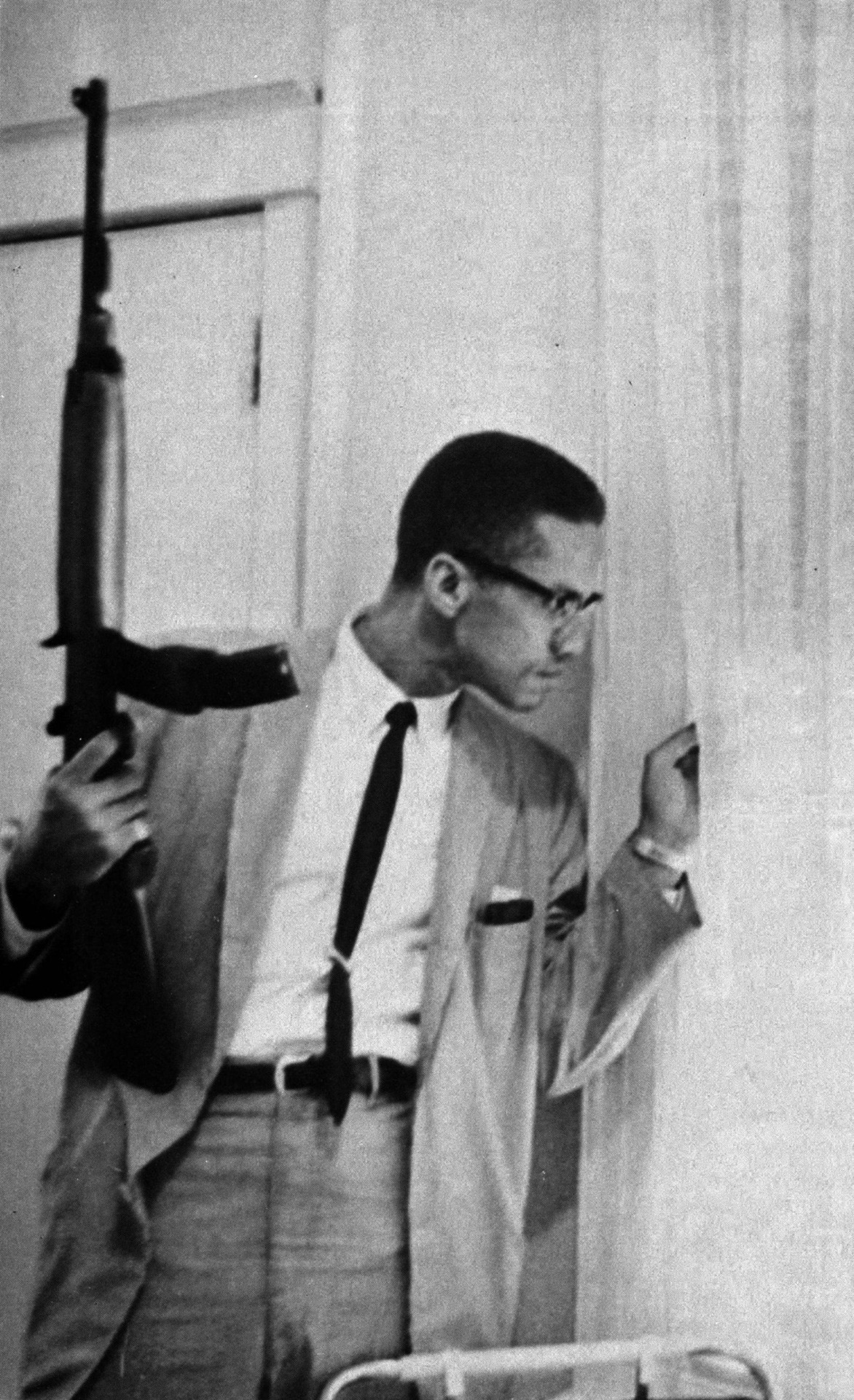American born Minister and Civil Rights activist, Malcolm X peeks out his window with an A-1 Carbine  rifle amidst death threats and violence he constantly received.