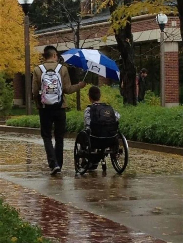 Guy holds his umbrella for this wheelchaired man to protect him from the rain.