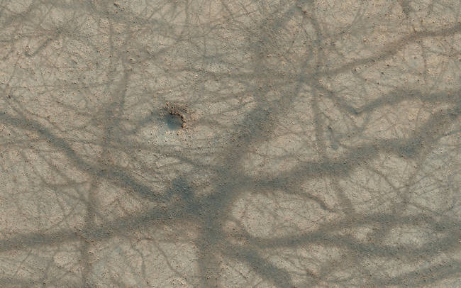 A small impact crater, pitted knobs, and a criss-cross mesh of dust devil trails across the martian surface.