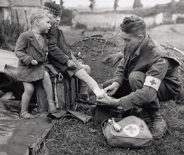 A medic bandages the injured foot of a child while his younger brother looks on. World War II, 1944