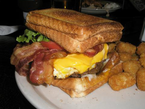 A burger topped with five slices of bacon, four slices of cheese, two fried eggs, mayo, lettuce, tomato, and onion between two grilled cheese sandwiches