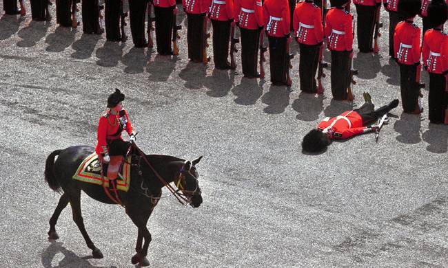 A foot guard passes out as Queen Elizabeth II rides past during a parade. [1970]