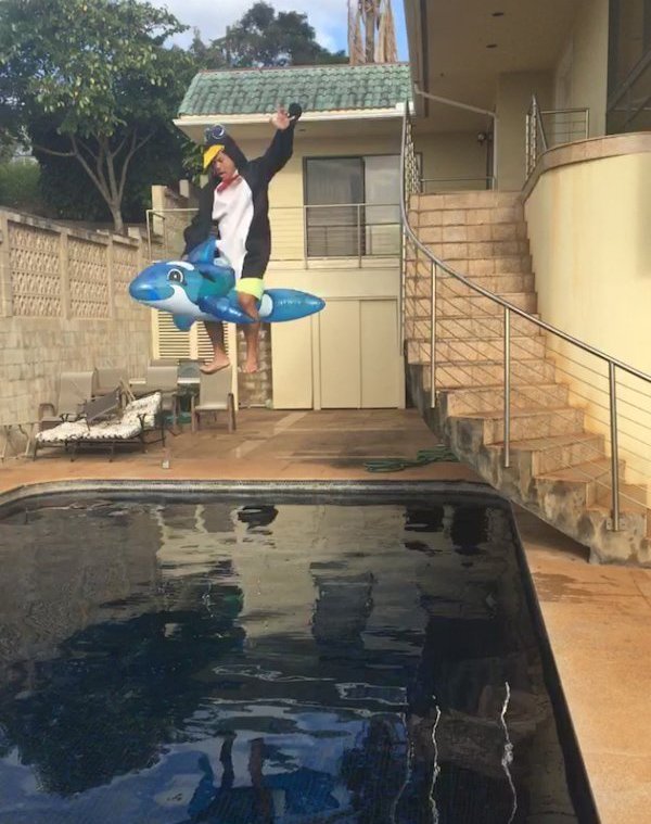 Man wearing silly pajamas and jumping off the stairs into a pool on an inflatable dolphin toy.