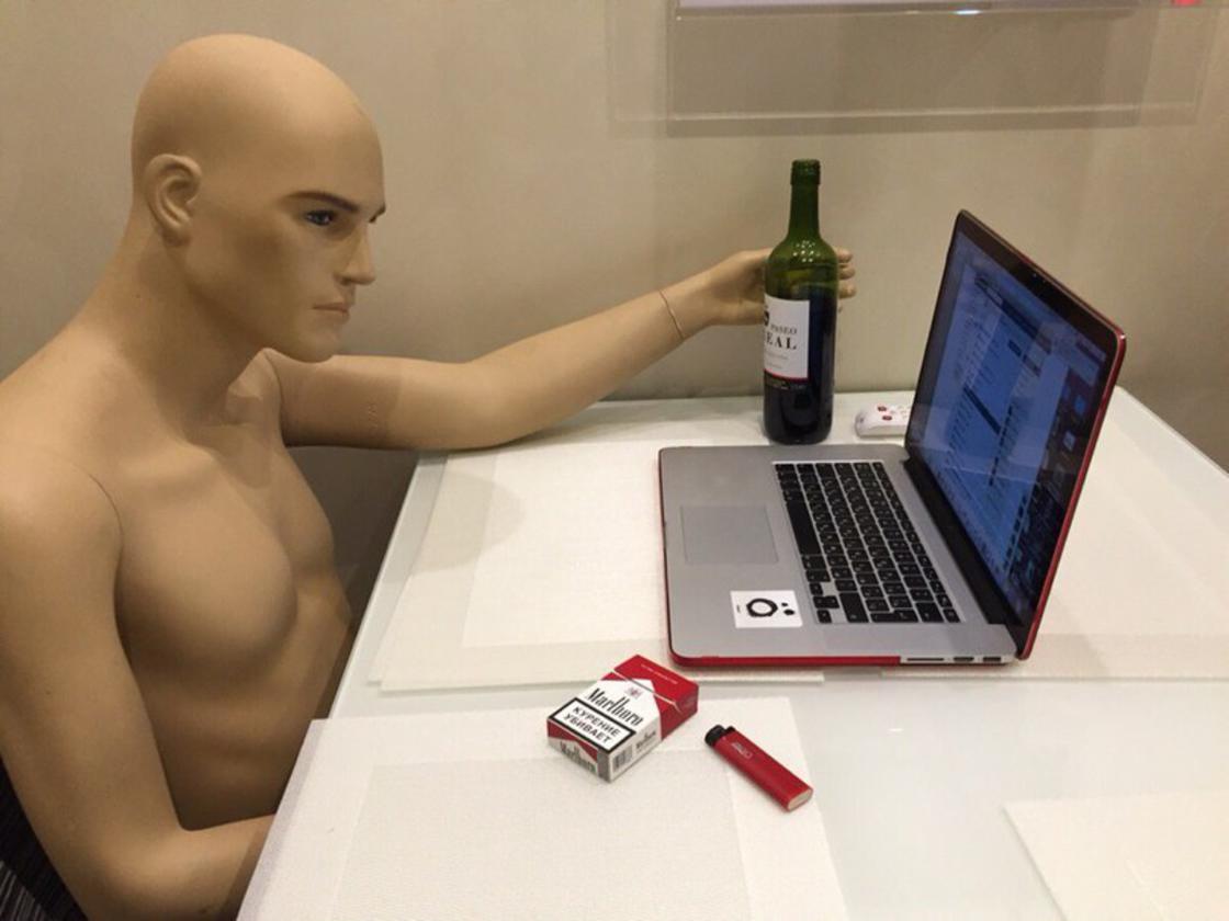 Mannequin at a computer with cigarettes and wine, with one hand under the table, doing who knows what.