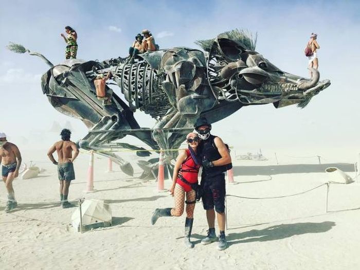 Surreal scene of couple taking picture in burning man in front of warthog sculpture.