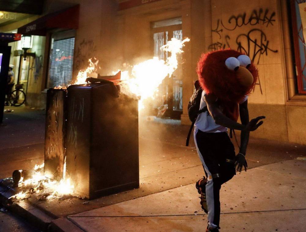 guy wearing big elmo head costume running away from trash cans on fire