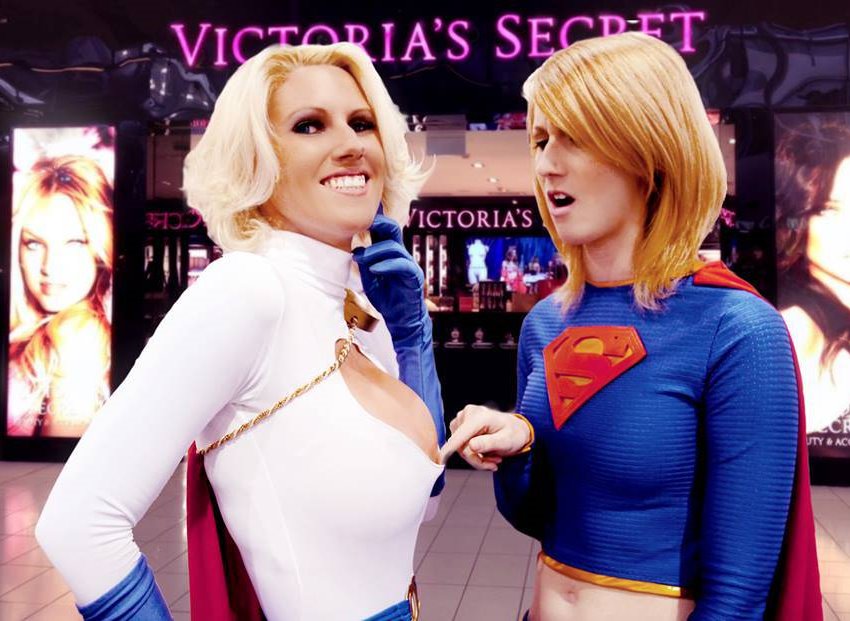 Two women cosplaying as SuperMan comparing breasts sizes. One woman has breast envy