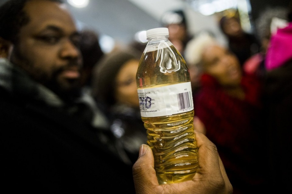 A pastor holds up a bottle of Flint water during protests outside of Michigan Gov. Rick Snyder’s office Thursday. The city’s been sending notices for past-due water bills even though the water has been poisoned with lead
