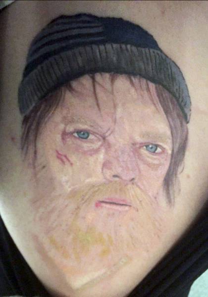 The tattoo shows shows the character with a scraggly beard, beanie hat and scratches on his face.