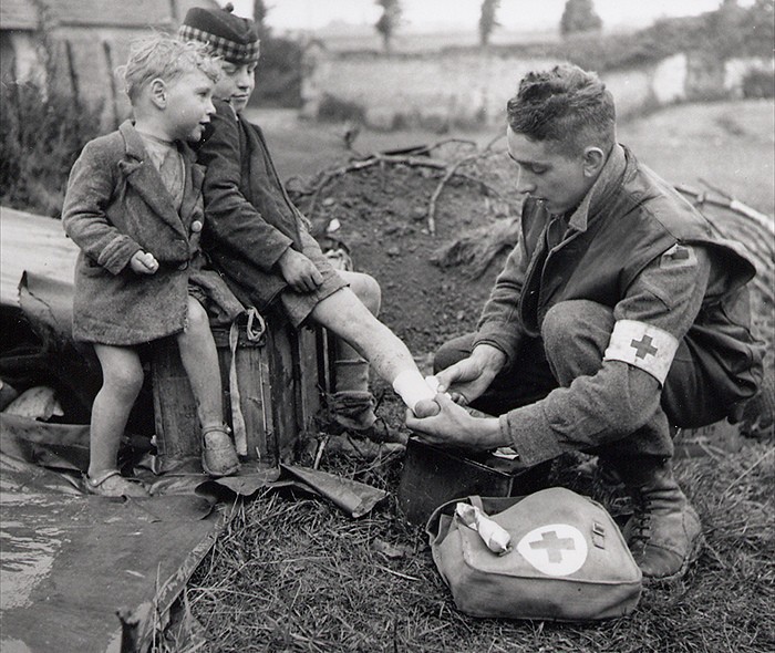 A medic bandages the injured foot of a child while his younger brother looks on. [World War II, 1944]