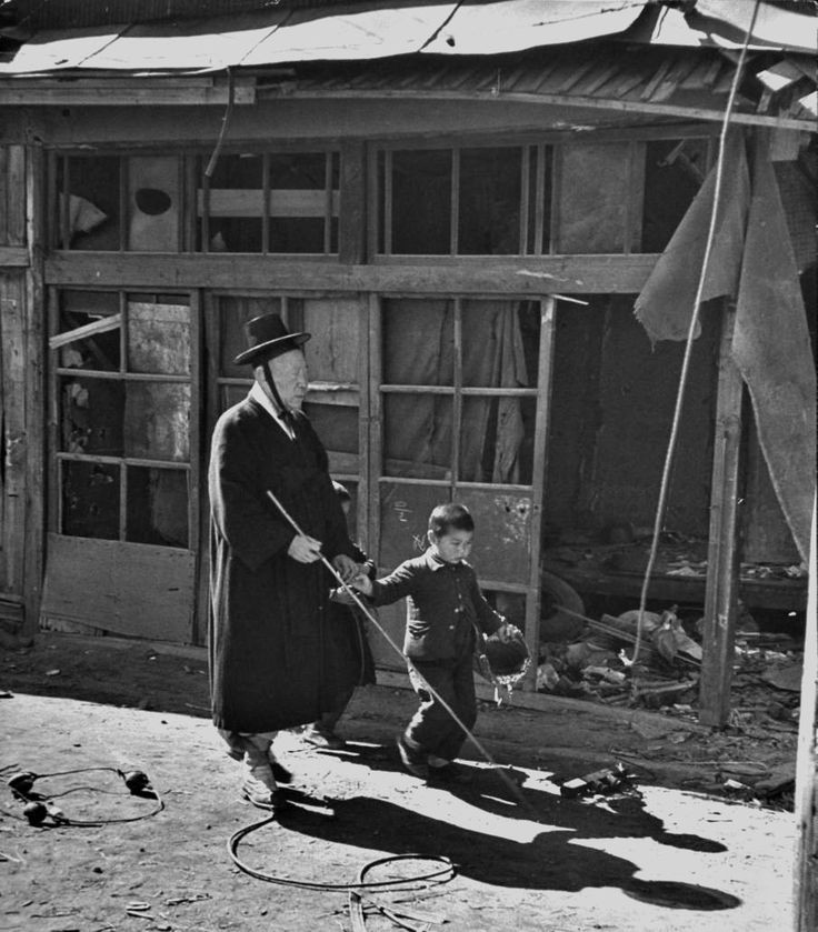 A boy leads an old blind man through war-torn streets. [Korean War, c, 1951]