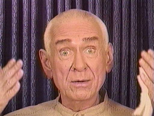Old photo of cult leader Marshall Applewhite gesturing with both hands with purple curtains in the background