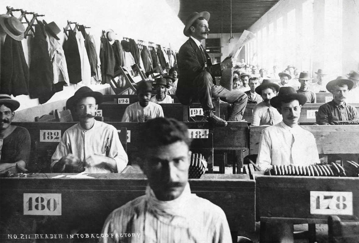 Tobacco factory Lectors, 1910.

The practice of reading aloud while others listen intently as they engage in manual labor has a long and distinguished tradition through out the Caribbean in the practice of cigar manufacture. Because the job of rolling cigar after cigar could become monotonous, the workers wanted something to occupy and stimulate the mind. Thus arose the tradition of lectors, who sat perched on an elevated platform in the cigar factory, reading to the workers. It started in Cuba and was brought to the United States more particularly to Key West in 1865 when thousands of Cuban cigar workers emigrated to Florida to escape Spanish oppression.
Historically, lectors or readers in a cigar factory entertained workers by reading books or newspapers aloud, often left-wing publications, paid for by unions or by workers pooling their money. The workers would each give 25 to 50 cents of their weekly salary to elect a fellow workman to act as “the reader” in which he would read aloud not just only newspapers, but even classical works of literature such as Tolstoy or Dickens.
The readers, elected by their peers, were actually marvelous actors and would not simply read the book but literally act out the scenes in a dramatic fashion upon a podium set up in the middle of the factory.
Workers were both generous and ruthless to the lectors, depending on the performance. If they enjoyed the day’s reading, they would loudly tap their chavetas on their cutting boards as a form of applause. On the other end of the spectrum, workers could vocalize their unhappiness with a particular reading. Since the lector was paid by the workers, he or she took cues from them. The lector committee, not the lector, chose the materials to be read. This mattered very little to the factory owners. The lectors were forced out of the factories when what they were reading was deemed too radical. This caused widespread strikes and work slowdowns.