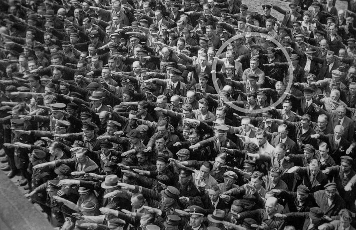 A lone man refusing to do the Nazi salute, 1936.
The photo was taken at the launch of a German army vessel in 1936, during a ceremony that was attended by Adolf Hitler himself. Within the picture a lone man stood with arms crossed as hundreds of men and women around him held up their arms in salute and allegiance to the Nazi Party and its leader, Adolph Hitler. Everyone in attendance is showing their undying support for Der Fuhrer by throwing out their very best “Sieg Heil.” August Landmesser, grimacing with arms crossed, stood strong and defiant as he showed his disapproval by not displaying support for the Nazi Party. What made this photo and Landmesser’s defiance unique is that it represented the protest of one man, in its most sincere and pure form. The source of Landmesser’s protest, like many great tragedies, starts with a love story.
The story of August Landmesser’s anti-gesture begins, ironically enough, with the Nazi Party. Believing that having the right connections would help land him a job in the pulseless economy, Landmesser joined the Nazi Party in 1931. Little did he know that his heart would soon ruin any progress that his superficial political affiliation might have made. In 1934, Landmesser met Irma Eckler, a Jewish woman, and the two fell deeply in love. Their engagement a year later got him expelled from the party, and their marriage application was denied under the newly enacted racial Nuremberg Laws. They had a baby girl, Ingrid, in October of the same year, and two years later in 1937, the family made a failed attempt to flee to Denmark, where they were apprehended at the border. August was arrested and charged for “dishonoring the race” under Nazi racial law. He argued that neither he nor Eckler knew that she was fully Jewish, and was acquitted on 27 May 1938 for lack of evidence, with the warning that a repeat offense would result in a multi-year prison sentence.
The couple publicly continued their relationship and a month later August Landmesser would be arrested again and sentenced to hard labor for two years in a concentration camp. He would never see his beloved wife again. Eckler was detained by the Gestapo and held at the prison Fuhlsbüttel, where she gave birth to a second daughter Irene. Their children were initially taken to the city orphanage. Ingrid was later allowed to live with her maternal grandmother; Irene went to the home of foster parents in 1941. Later, after her grandmother’s death in 1953, Ingrid was also placed with foster parents. A few letters came from Irma Eckler until January 1942. It is believed that she was taken to the so-called Bernburg Euthanasia Centre in February 1942, where she was among the 14,000 killed. In the course of post-war documentation, in 1949 she was pronounced legally dead, with a date of 28 April 1942.
The first and only photo of the family, June 1938. Although it was forbidden for them to meet, they appeared together in public and put themselves at exceptional risk. The first and only photo of the family, June 1938. Although it was forbidden for them to meet, they appeared together in public and put themselves at exceptional risk. August would be released in 1941 and began work as a foreman. Two years later, as the German army became increasingly mired by its desperate circumstances, Landmesser would be drafted into a penal infantry along with thousands of other men. He would go missing in Croatia where it is presumed he died, six months before Germany would officially surrender. His body was never recovered. Like Eckler, he was declared legally dead in 1949.