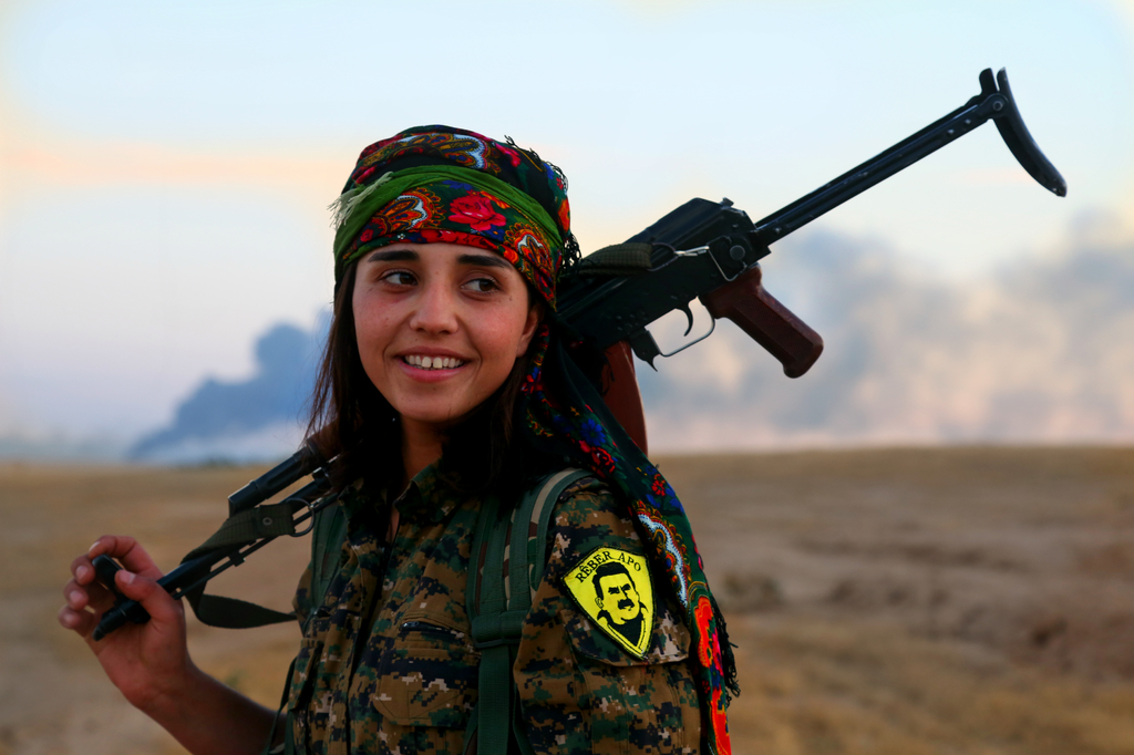 A Kurdish YPJ fighter with smoke behind her rising from an ISIS held area