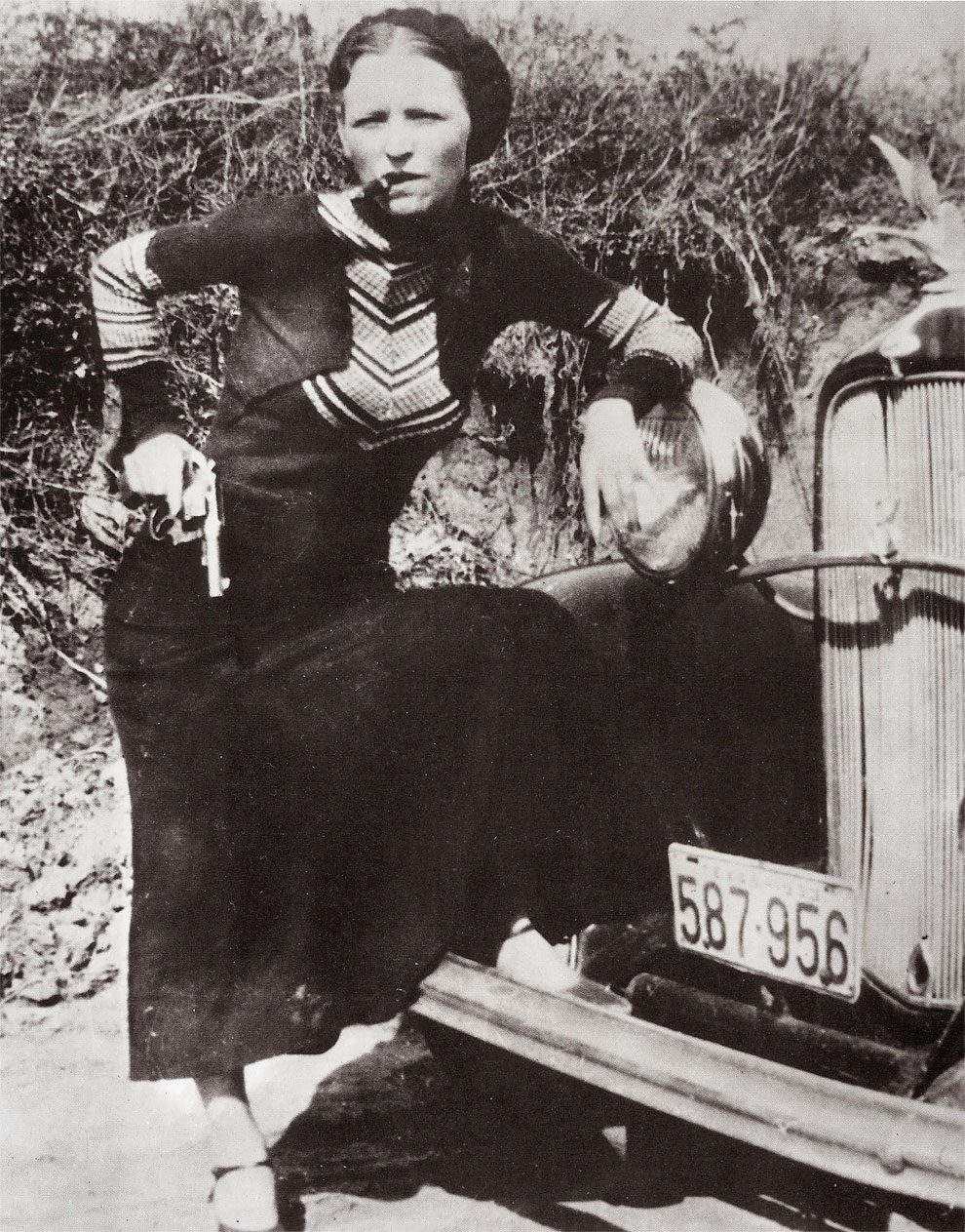 Full-length portrait of American criminal Bonnie Parker (1910 – 1934) smoking a cigar while leaning on the front fender of a car and holding a pistol on April 17, 1933