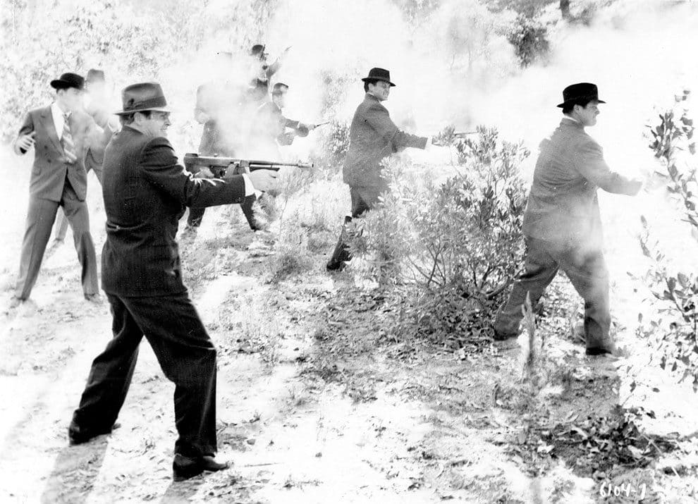 A group of men in suits and hats are obscured by the smoke from the guns, including Thompson submachine guns, shotguns, and revolvers, that they are firing in a shrubland, USA, 1930s