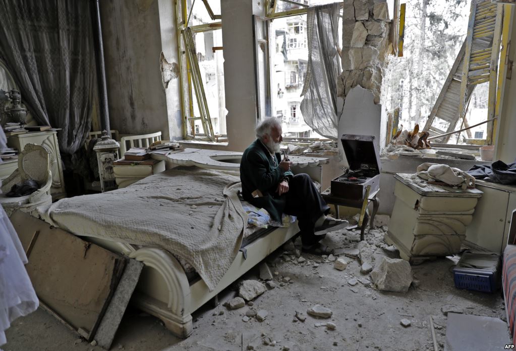 70-year-old man sits in his destroyed bedroom, smoking a pipe and listening to music on his record player, in Aleppo, Syria