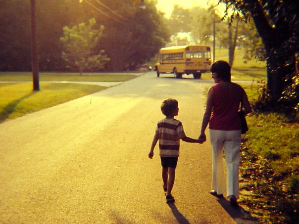 Kid walking home with mom, holding hands, and school bus drives off in the distance.
