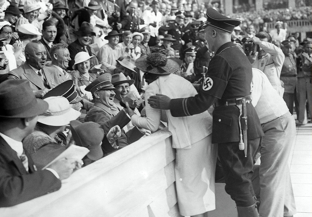 Hitler Reacts to Kiss from Excited American Woman at the Berlin Olympics, August 15, 1936.

Shortly before the finish of the men’s 1500 meters free-style swimming, a woman in a red hat, whom Black Guards repeatedly prevented from photographing Hitler at close range, broke the cordon during the excitement of the finish of the race, shook Hitler by the hand and then kissed him, while the crowd of 20,000 rocked with laughter. Hitler, who was in high spirits, joined in the fun, clapping his hands as the woman returned triumphantly to her seat.
The woman, Carla De Vries, was a 40-year old American traveling in Europe. Her brother-in-law was quoted in that “She wanted to meet Hitler but I’m surprised at the way she did it”. Her rationale from her own words? “Why? I simply embraced him because he appeared so friendly and gracious. People sitting near Der Fuehrer’s box began to cheer and applaud so loudly that I ran back to my husband and told him we had better leave. I don’t know why I did it. Certainly I hadn’t planned such a thing. It’s just that I’m a woman of impulses, I guess. It happened when I went down to take Hitler’s picture with my small movie camera. Hitler was leaning forward, smiling, and he seemed so friendly that I just stepped up and asked for his autograph, which he wrote on my swimming ticket. He kept on smiling and so I kissed him”.
For their failure to halt the woman, several of Hitler’s hulking Schutzstaffel guards were dismissed in disgrace, several more were demoted in rank.