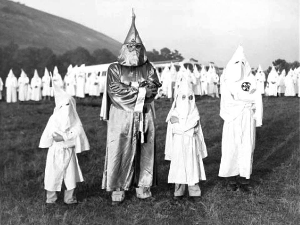 Two children wearing Ku Klux Klan robes and hoods stand on either side of Dr. Samuel Green, Ku Klux Klan Imperial Wizard, at an initiation ceremony. Atlanta, Georgia. July 24, 1948