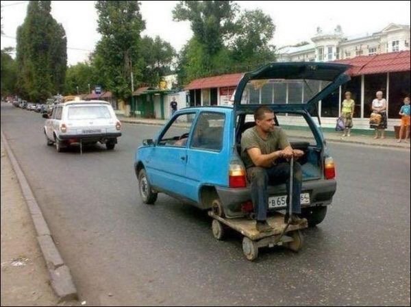 Man on a car in russia that is missing a wheel and a wagon is under where the wheel goes.
