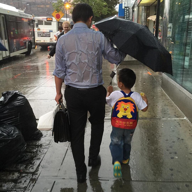 A father covers his son with the umbrella and is soaked by the rain.