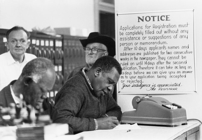 Black citizens fill out voter registration forms at the Courthouse under a sign warning them the applicants’ names and addresses will be published in the newspapers. Hattiesburg, Forrest County, Mississippi, United States. 1964
