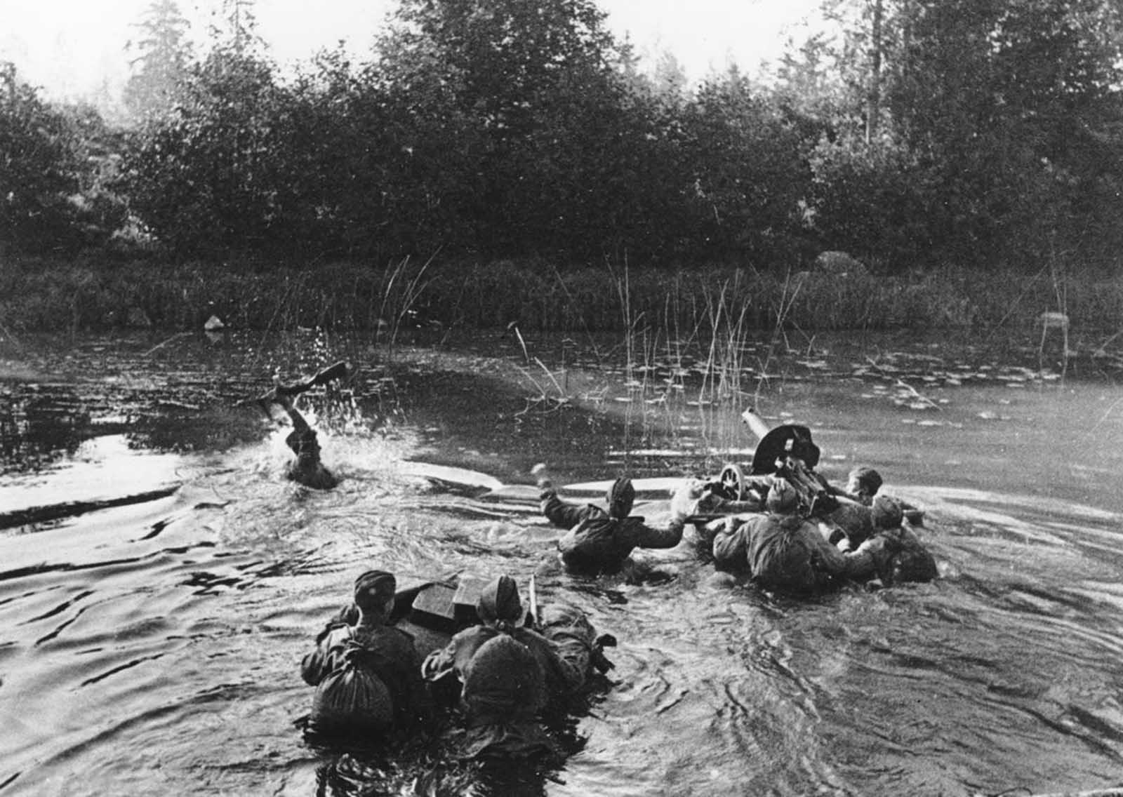 A Soviet machine gun crew crosses a river along the second Baltic front, in January of 1945. The soldier on the left is holding his rifle overhead while his comrades push a floating device with a Maxim machine gun, followed by two men with several supply boxes.