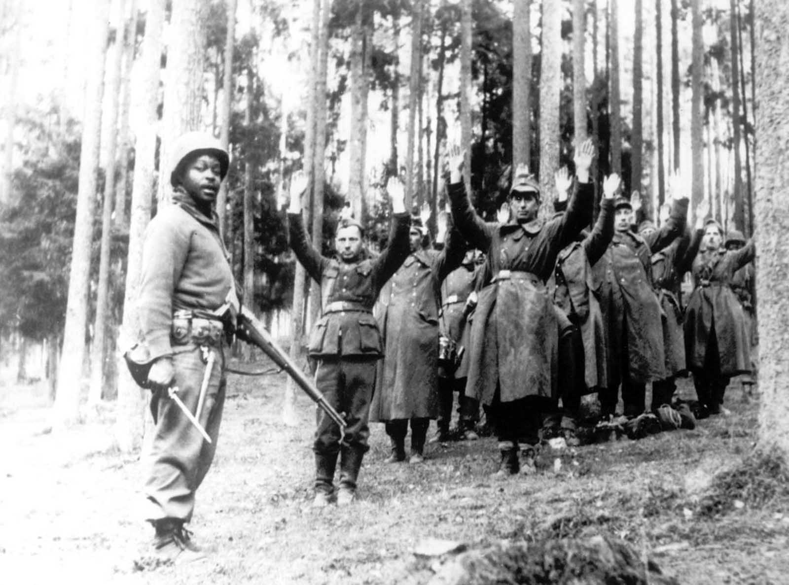 An American soldier of the 12th Armored Division stands guard over a group of German soldiers, captured in April 1945, in a forest at an unknown location in Germany.