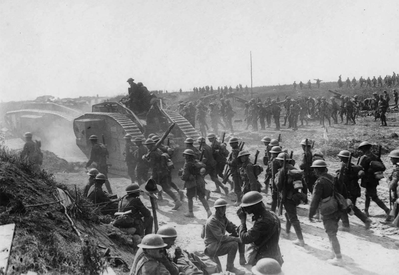Allied advance on Bapaume, France, ca. 1917. Two tanks are moving towards the left, followed by troops. In the foreground some soldiers are sitting and standing at the roadside. One of them appears to be having a drink. Beside the men is what appears to be a rough wooden cross with an Australian or New Zealand service hat on it. In the background other troops are advancing, moving field guns and mortars