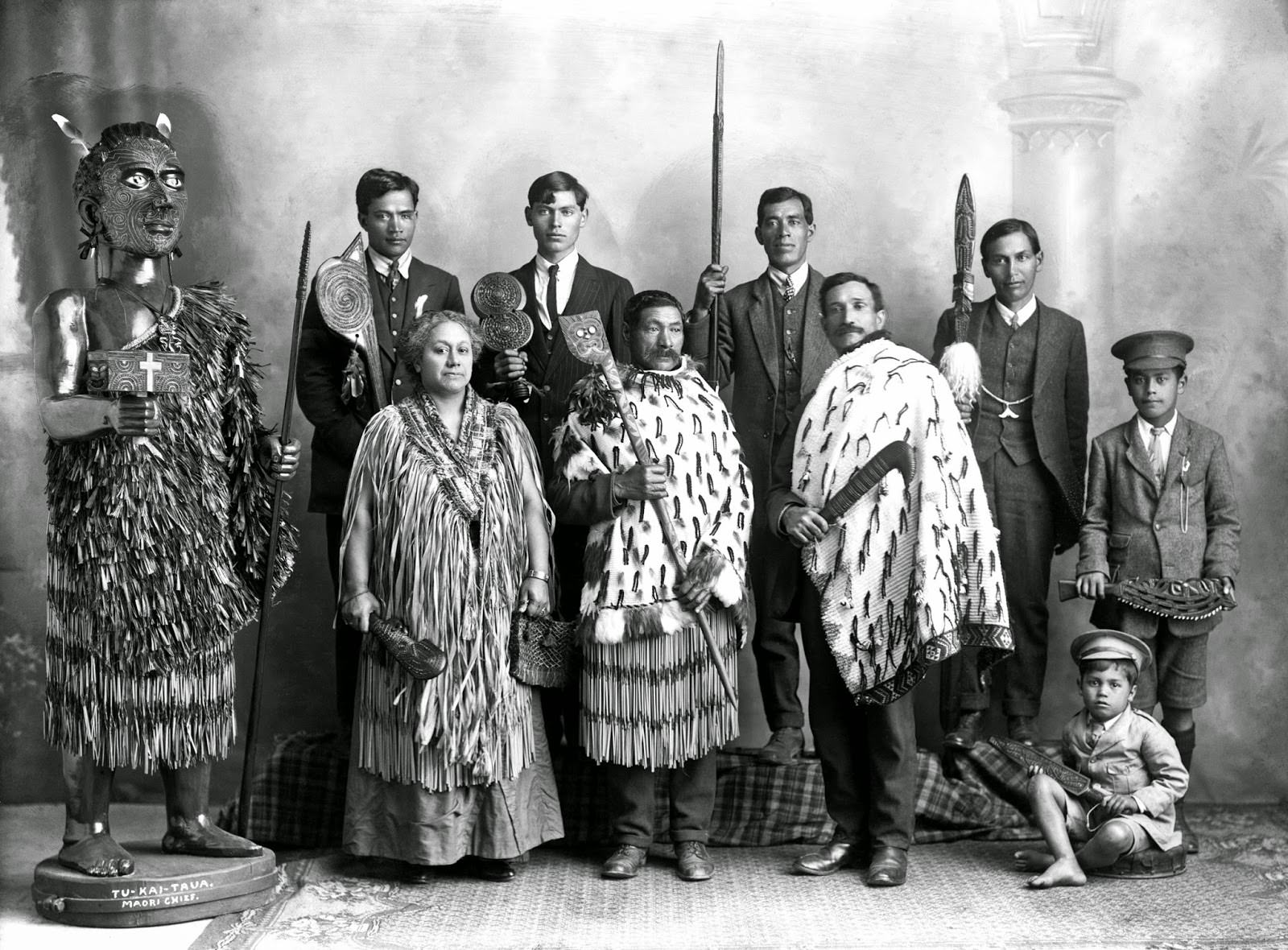 A Maori family standing alongside a carved figure of the Maori chief Tu-Kai-Taua and each person holding a carved wooden Maori weapon in New Zealand in 1915.