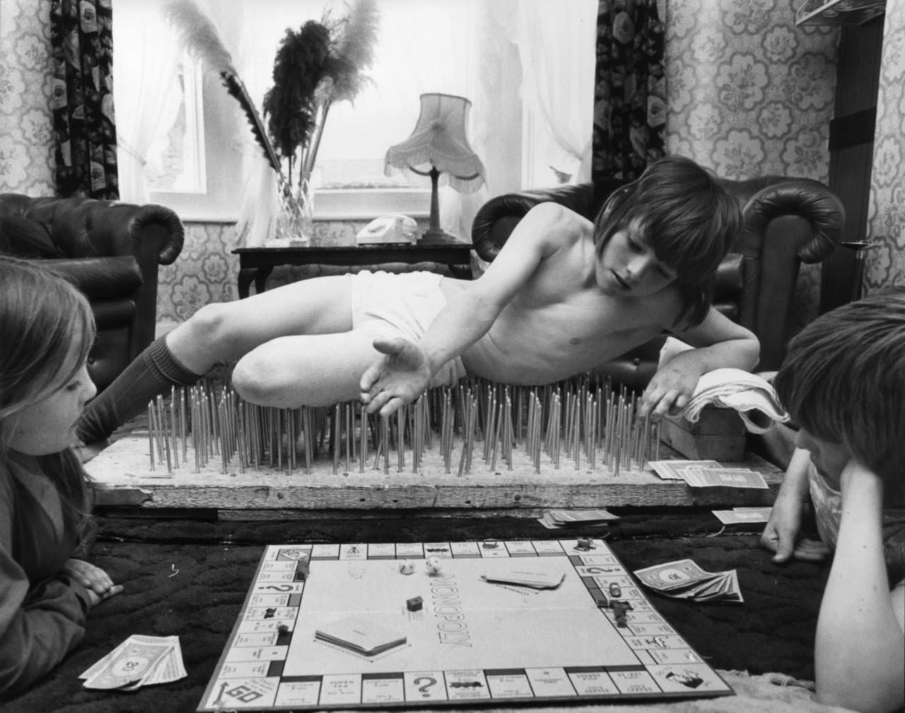 An 8 year old boy named Mark lies on a bed of nails while playing monopoly in London, England in 1976.