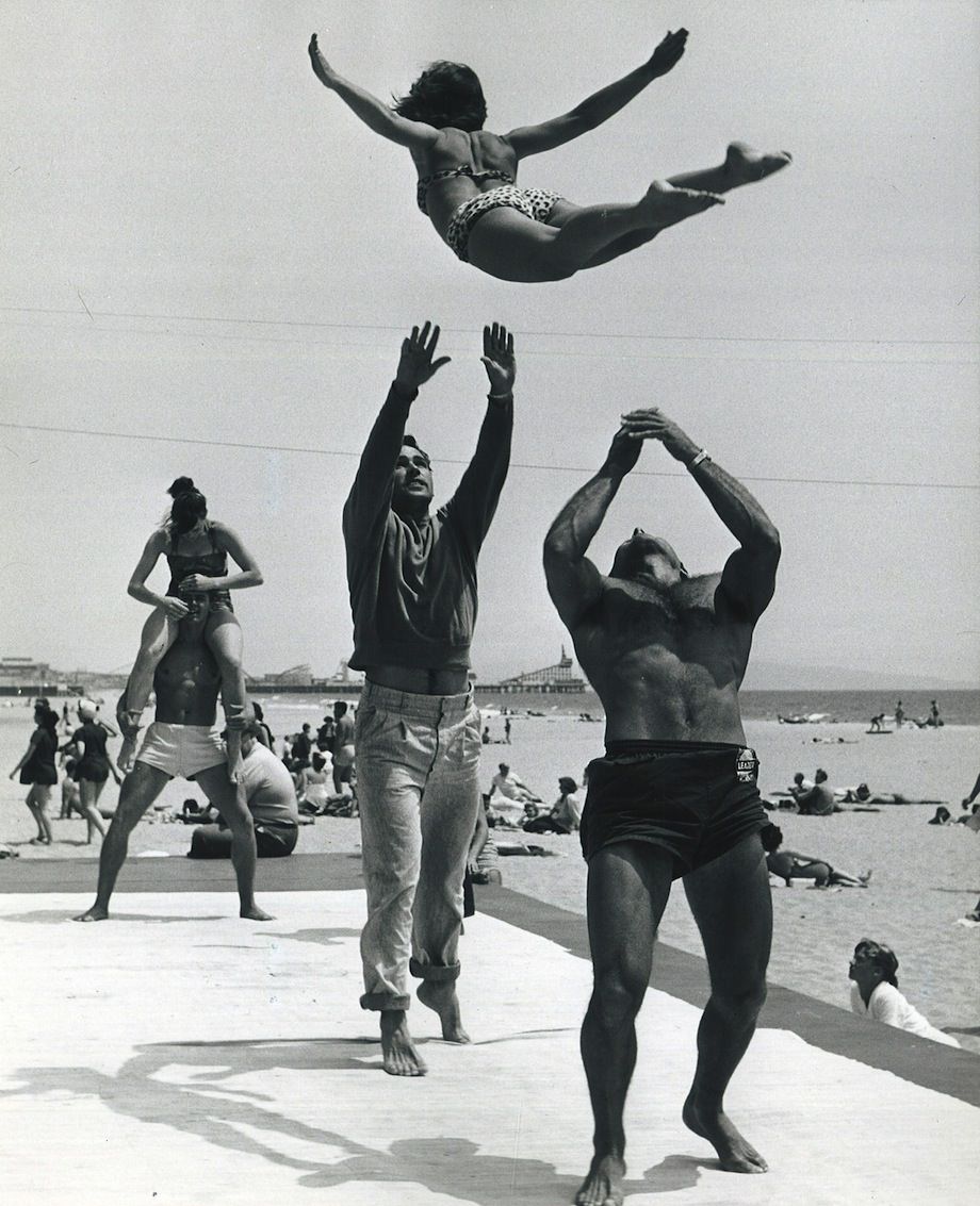 Beach weight lifters do some acrobats with some woman for a photographer in CA, US in 1957.