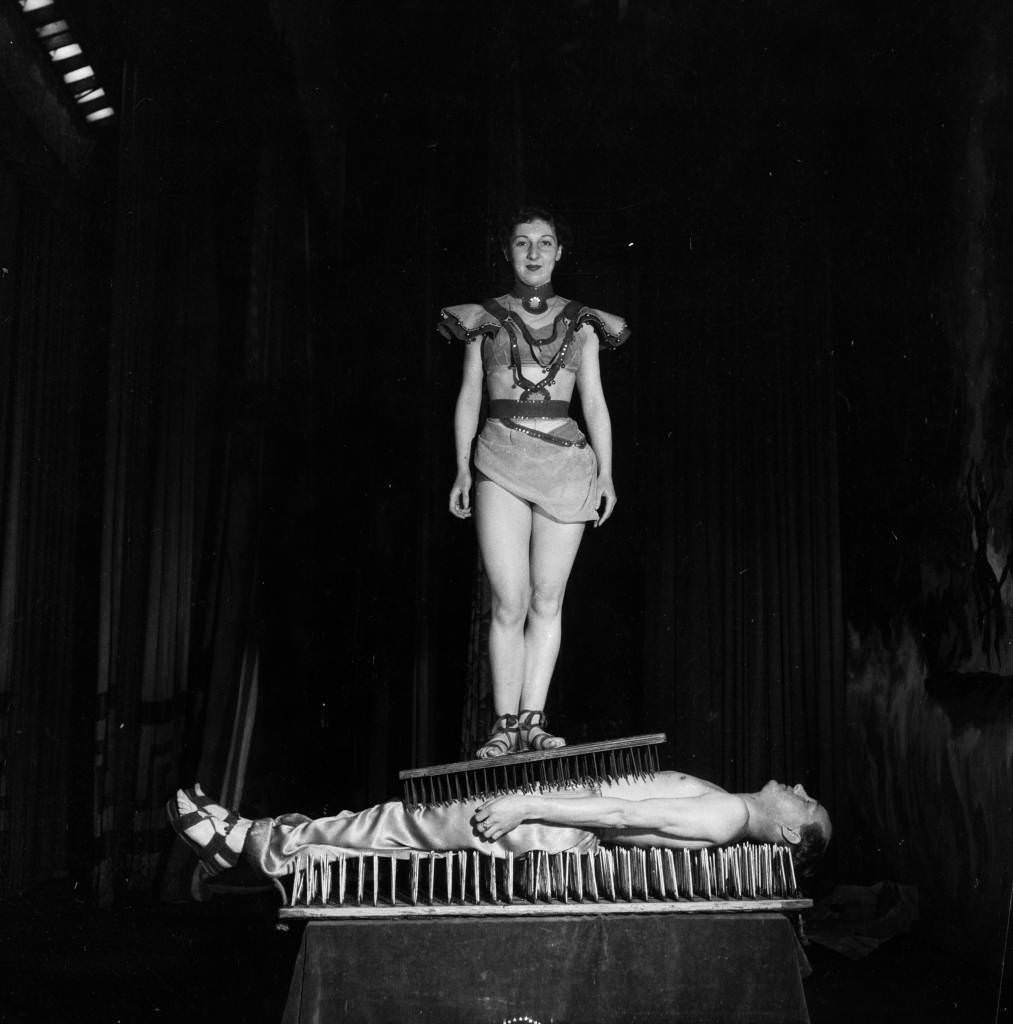 A Fakir has his assistant stand on a board of nails while he lies on another board of nails for a show in England in 1955.