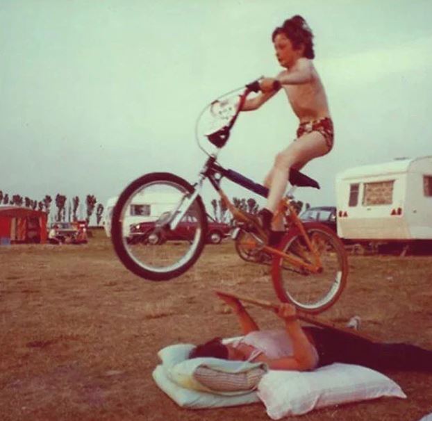 Vintage photo of a kid riding a bmx bike off a ramp that's just a woman laying on the ground with a board