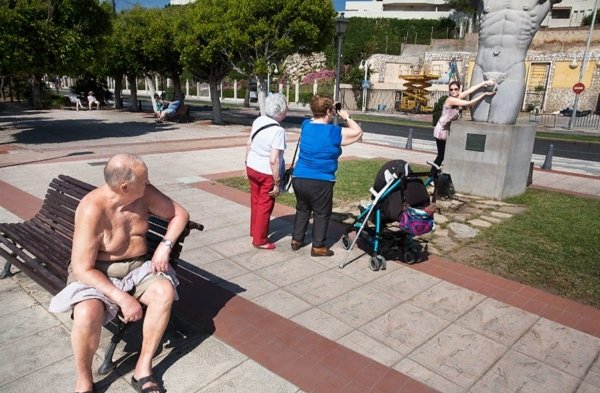 Girl grabbing the genitals of a statue of a man while two old ladies take her picture and an old man with her shirt off sitting on a bench looks on