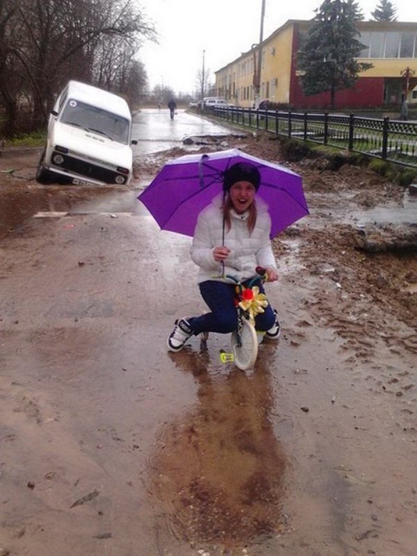 Woman with a purple umbrella riding a tiny bicycle in the rain with a truck crashed into a massive pothole behind her