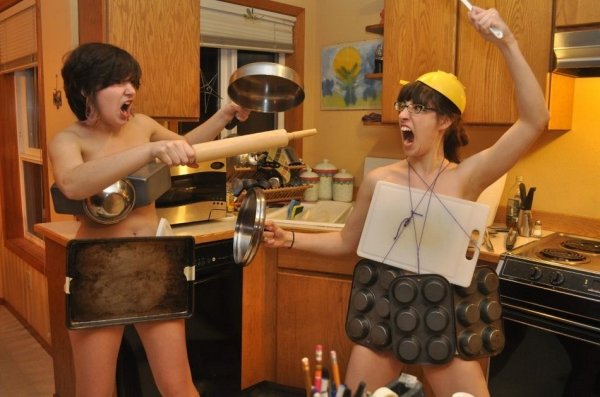 Two women pretending to fight while wearing nothing but cookie sheets, cutting boards and muffin pans