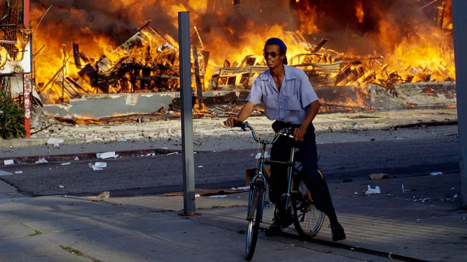 Man rides his bikes while flames burst all around during the 1992 Los Angeles Riots