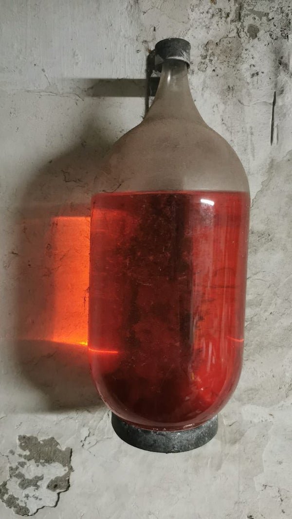 Found during cellar renovation in Belgium. A sealed container mounted on the wall with a mysterious red liquid inside.

A: Carbon tet fire extinguisher. Liquid is likely to be very toxic. Should be removed and disposed of by a specialist.