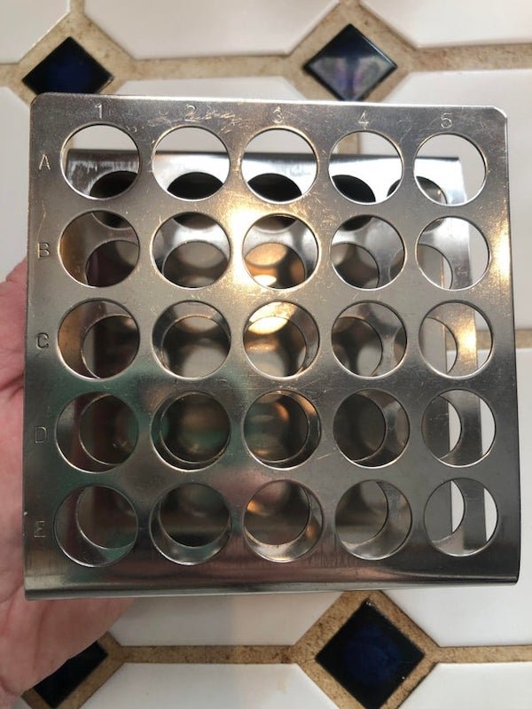 What is this 6 inch by 6 inch square stainless steel item with 25 holes in it labeled 1-5 across the top and A-E down the left side? Found in a thrift store in Buffalo, NY.

A: An old school test tube rack.