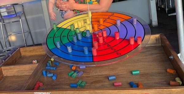 What game is this? You roll dice with colors and numbers and place the colored pegs on the wheel accordingly while trying maintain balance.

A: It’s Dingo Disc